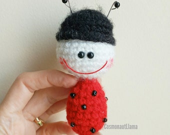 Puede incluir: Un juguete de mariquita de crochet con un sombrero negro, una cara blanca, un cuerpo rojo y puntos negros.