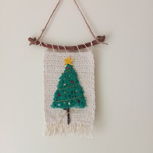 May include: A crocheted Christmas tree wall hanging with a green tree, red and green baubles, a yellow star, and a brown branch at the top. The hanging is made of natural fibre and has a fringe at the bottom.