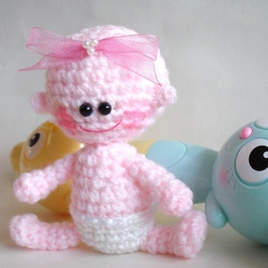 May include: A pink crocheted baby doll with a pink bow and white diaper. The doll has black eyes and a smiling face.