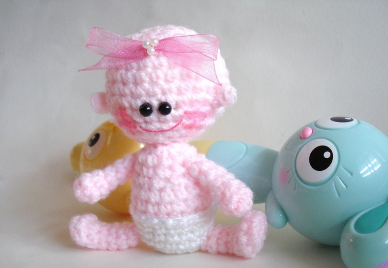 May include: A pink crocheted baby doll with a pink bow and white diaper. The doll has black eyes and a smiling face.