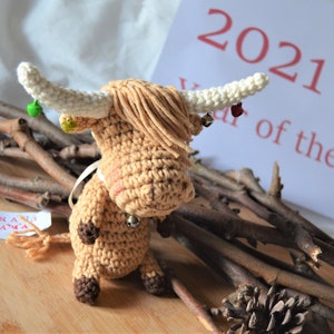 Puede incluir: Un adorno de vaca de las tierras altas de crochet marrón y blanco con cascabeles, una manzana verde y una cinta blanca. El adorno está sobre una superficie de madera con ramas marrones y una piña. El fondo es una superficie blanca con una tarjeta blanca que dice "2021 Año del Buey".