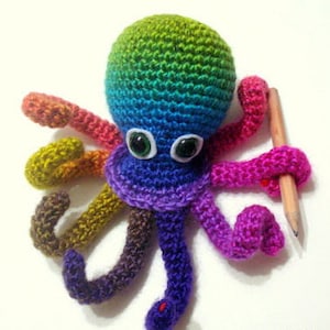May include: A rainbow crocheted octopus with white googly eyes holding a pencil. The octopus is made with different colored yarn, including green, blue, purple, pink, and brown.