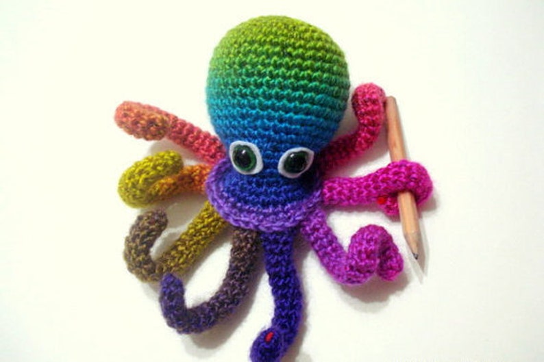 May include: A rainbow crocheted octopus with white googly eyes holding a pencil. The octopus is made with different colored yarn, including green, blue, purple, pink, and brown.