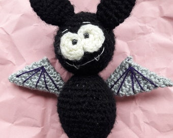 May include: A crocheted black bat with gray wings and white eyes. The bat has a stitched smile and is sitting on a pink background.