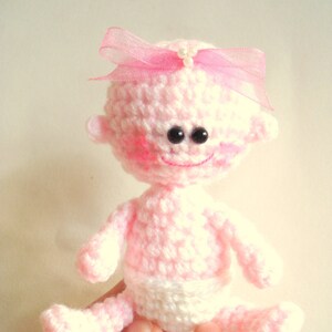 May include: A pink crocheted baby doll with a pink bow and black eyes. The doll is wearing a white crocheted diaper.