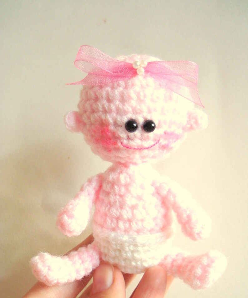 May include: A pink crocheted baby doll with a pink bow and black eyes. The doll is wearing a white crocheted diaper.
