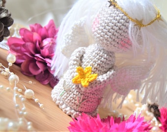 Könnte beinhalten: Eine gehäkelte Engelpuppe aus weißem Garn, die eine gelbe Blume in der Hand hält. Der Engel trägt ein goldenes Stirnband mit Sternchen. Die Puppe steht auf einer Holzoberfläche mit rosa Blumen und einer weißen Perlenkette.