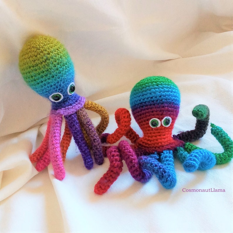 May include: Two crocheted rainbow octopus plushies with googly eyes. The octopus on the left has pink, purple, brown, and orange tentacles. The octopus on the right has red, purple, blue, green, and black tentacles.