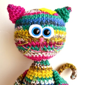 Könnte beinhalten: Ein farbenfrohes Häkeltier in Form einer Katze mit großen blauen Augen. Die Katze ist aus verschiedenen Garnfarben gefertigt, darunter Pink, Grün, Gelb und Braun.