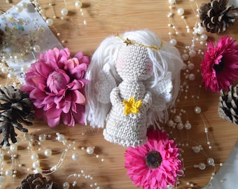 Könnte beinhalten: Eine gehäkelte Engelpuppe mit weißen Garnhaaren, einem goldenen Heiligenschein und einer gelben Blume in den Händen. Die Puppe ist umgeben von einer weißen Perlenkette, rosa Blumen und Tannenzapfen auf einer Holzoberfläche.