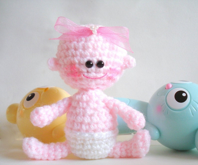 May include: A pink crocheted baby doll with a pink bow and black eyes. The doll is wearing a white crocheted diaper.