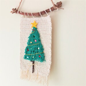 May include: A crocheted Christmas tree wall hanging with a green tree, a yellow star, and red and white ornaments. The tree is attached to a natural branch with twine.