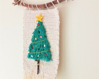 Op de afbeelding: Een gehaakte kerstboomwanddecoratie met een groene boom, een gele ster en rode en witte ornamenten. De boom is met touw aan een natuurlijke tak bevestigd.