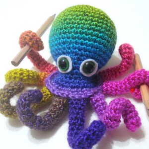 May include: A colorful crocheted octopus holding two pencils. The octopus is made with rainbow colored yarn and has large green eyes.