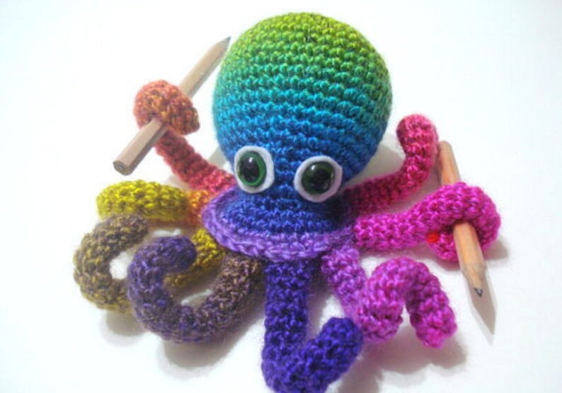 May include: A colorful crocheted octopus holding two pencils. The octopus is made with rainbow colored yarn and has large green eyes.