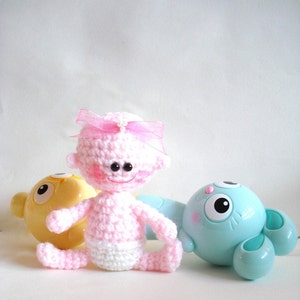 May include: A pink crocheted baby doll with a pink bow and a white diaper. The doll is sitting next to a yellow and a blue toy.