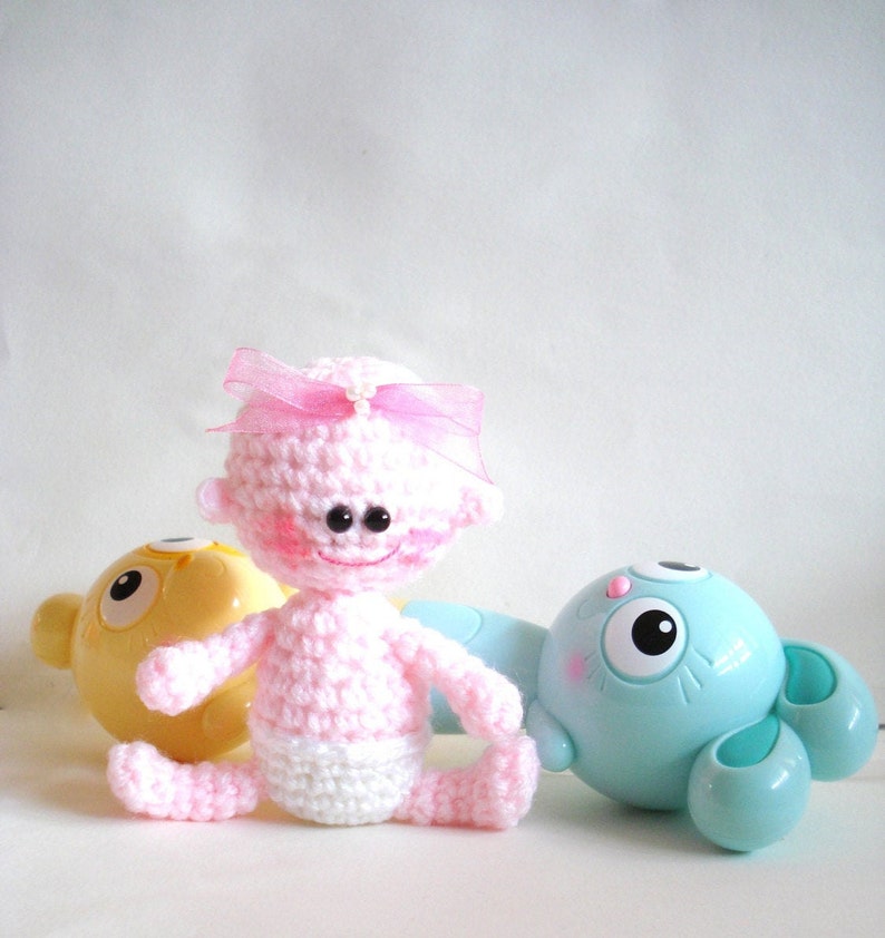 May include: A pink crocheted baby doll with a pink bow and a white diaper. The doll is sitting next to a yellow and a blue toy.