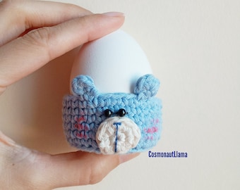 May include: A blue crocheted egg cup shaped like a bear's face. The egg cup has pink accents and black eyes. An egg is sitting inside the cup.