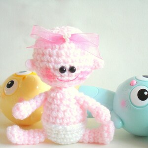 May include: A pink crocheted baby doll with a pink bow and white diaper. The doll has large black eyes and a smiling face. The doll is sitting on a white surface with a yellow and blue toy in the background.