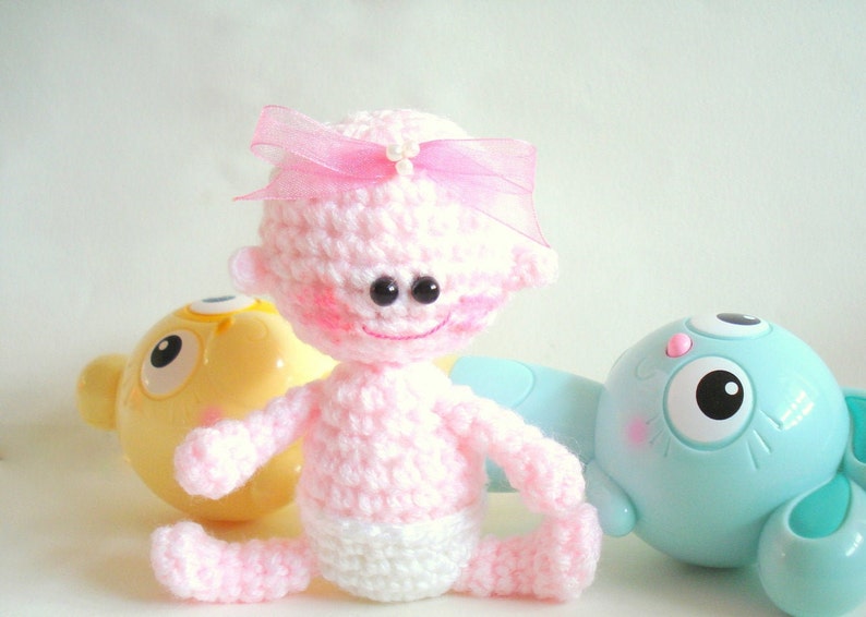 May include: A pink crocheted baby doll with a pink bow and white diaper. The doll has large black eyes and a smiling face. The doll is sitting on a white surface with a yellow and blue toy in the background.