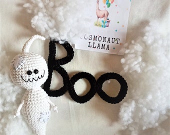May include: A white crocheted ghost with a stitched-on smile and eyes, next to a black crocheted word "BOO" on a white background.