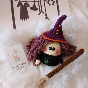 Puede incluir: Una muñeca de bruja de crochet morado y naranja con una larga escoba de crochet marrón. La muñeca lleva un vestido negro y un sombrero morado con ribete naranja y estrellas de colores. La muñeca está sentada sobre una superficie blanca y esponjosa.
