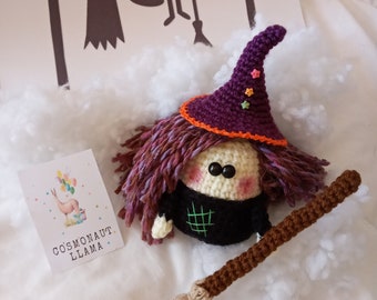 May include: A purple and orange crocheted witch doll with a long brown crocheted broom. The doll has a black dress and a purple hat with orange trim and colourful stars. The doll is sitting on a white fluffy surface.