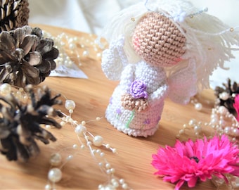 May include: A crocheted angel figurine with white yarn, a pink face, and a purple flower in its hand. The angel is standing on a wooden surface with white pearl beads and pink flowers.