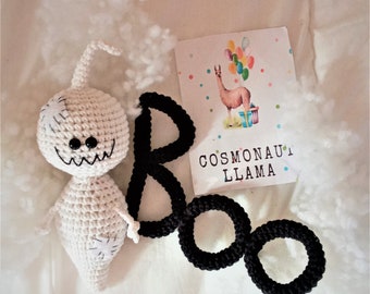 May include: A white crocheted ghost with black eyes and a stitched-on mouth, holding a black crocheted word "BOO" with a white background.