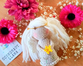 Könnte beinhalten: Eine gehäkelte Engelpuppe mit weißen Haaren, einer goldenen Krone und einer gelben Blume. Die Puppe hält eine Blume und ist von rosa Blumen und weißen Perlen umgeben.