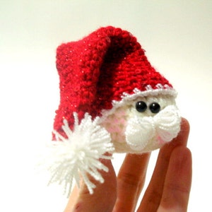 Op de afbeelding: Een klein gehaakt kerstman ornament met een rode muts en witte rand. Het ornament heeft zwarte ogen en een witte baard.