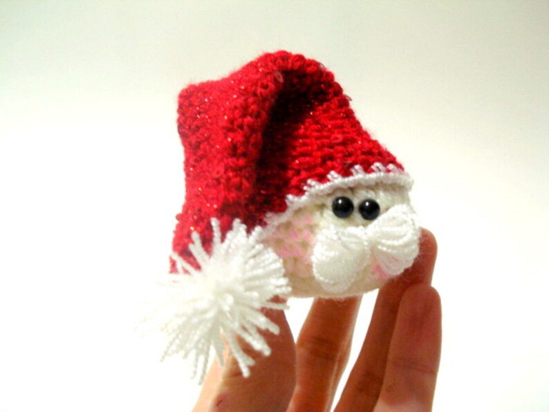 Op de afbeelding: Een klein gehaakt kerstman ornament met een rode muts en witte rand. Het ornament heeft zwarte ogen en een witte baard.