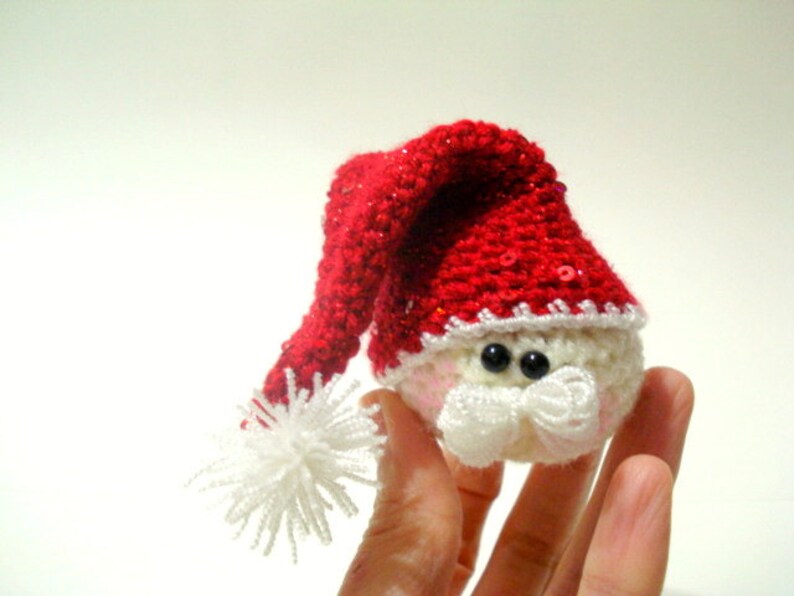 Op de afbeelding: Een klein gehaakt kerstman ornament met een rode muts, witte baard en zwarte ogen. Het ornament wordt in een hand gehouden.