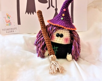 May include: A crocheted witch doll with purple and pink hair, a purple hat with stars, and a brown broom. The doll is sitting on a white surface.