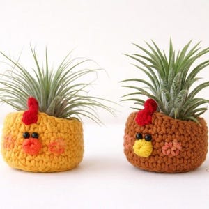 May include: Two crochet chicken planters, one yellow and one brown, each holding a green air plant. The planters have red combs, orange beaks, and black eyes. The yellow planter has orange cheeks, while the brown one has pink cheeks.