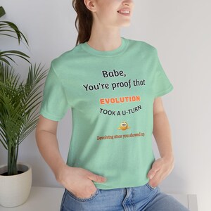 Può includere: Una maglietta verde chiaro con la scritta "Babe, You're proof that EVOLUTION TOOK A U-TURN" in nero e arancione. Sotto il testo c'è un personaggio dei cartoni animati. La maglietta è realizzata in un materiale morbido.