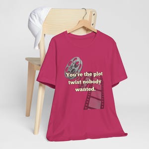 Puede incluir: Una camiseta magenta con un carrete de película y un gráfico de tira de película. El texto de la camiseta dice: "You're the plot twist nobody wanted." La camiseta está colgada en una percha de madera.