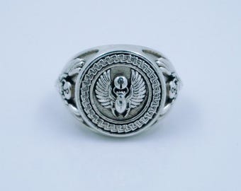 May include: Silver ring with a detailed winged scarab beetle design within a circular frame. The ring has an ornate, textured band and a raised central emblem. The design is reminiscent of ancient Egyptian jewellery. The ring is made of silver.