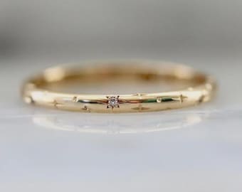May include: A gold-colored ring with a small diamond at the center. The ring is adorned with star-shaped engravings around the band. The ring is a delicate piece of jewelry.