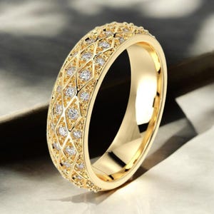 May include: A gold-colored ring with a diamond-shaped pattern. The ring is adorned with small, round, clear gemstones set within the diamond shapes. The band has a smooth, polished finish, and the ring appears to be made of gold.