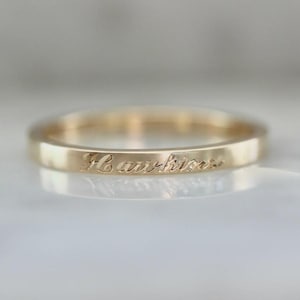 Puede incluir: Un anillo de banda de oro con la palabra "Hawkins" grabada. El anillo tiene un perfil plano y un acabado pulido. El anillo es probablemente de oro y se muestra sobre una superficie reflectante.