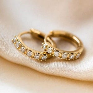 Op de afbeelding: Een paar gouden oorringen met een rij kleine, heldere edelstenen. De oorbellen liggen op een zachte, crèmekleurige stof, wat een luxe en elegante uitstraling creëert. De edelstenen schitteren en vangen het licht.