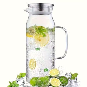 May include: A clear glass pitcher with a stainless steel lid and handle, filled with water, ice, lemon slices, and mint leaves. Limes, mint, and ice cubes are arranged around the base, suggesting a refreshing beverage. The pitcher has measurement markings.