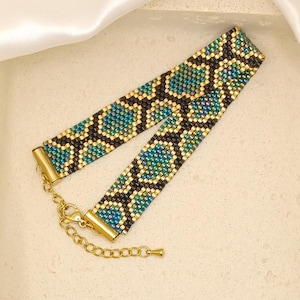 May include: A beaded bracelet with a geometric pattern in teal, gold, and black. The bracelet has a gold-colored clasp and chain for adjustable sizing. The design features a repeating pattern of shapes, creating a textured look. The bracelet is displayed on a neutral surface.