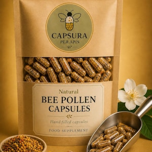 May include: A brown paper packet of natural bee pollen capsules, with the text "CAPSURA PER APIS". The capsules are displayed with a scoop and a small bowl of bee pollen. A white flower is in the background.