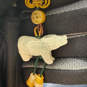 Puede incluir: Un colgante de jade con una figura de oso tallada en blanco y verde claro. El colgante está suspendido de un nudo dorado e incluye cuentas amarillas y naranjas. El oso tiene detalles tallados.