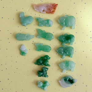 May include: A collection of small jade animal figurines in shades of green, white, and orange. The figurines include a rooster, pigs, dogs, and other animals. The background is a light yellow color with small dots.