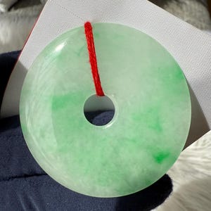 Könnte beinhalten: Ein runder, durchscheinender Jade-Anhänger mit einem zentralen Loch und einer roten Kordel. Der Jade weist verschiedene Grün- und Weißtöne auf, die einen marmorierten Effekt erzeugen. Der Anhänger wird vor einem weißen Hintergrund präsentiert.