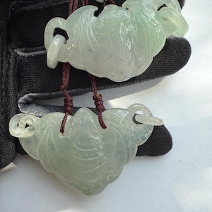 Puede incluir: Dos colgantes de jade con diseño de murciélago. Los colgantes son de color verde claro y translúcidos, con intrincadas tallas. Cuelgan de un cordón rojo oscuro y una pequeña cuenta verde.