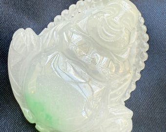 Op de afbeelding: Een doorschijnende jade carving van een zittende Boeddha figuur. De carving is voornamelijk wit met een vleugje groen en heeft ingewikkelde details. De Boeddha wordt afgebeeld in een ontspannen pose, met een serene uitdrukking. Donkerblauwe achtergrond.